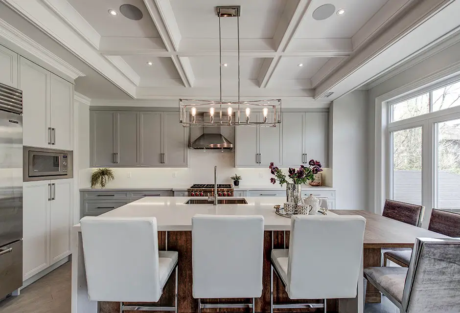 Luxurious white interior featuring an ornate coffered ceiling, decorative wall paneling, marble fireplace, and dark hardwood floors in an elegant open-concept living space.