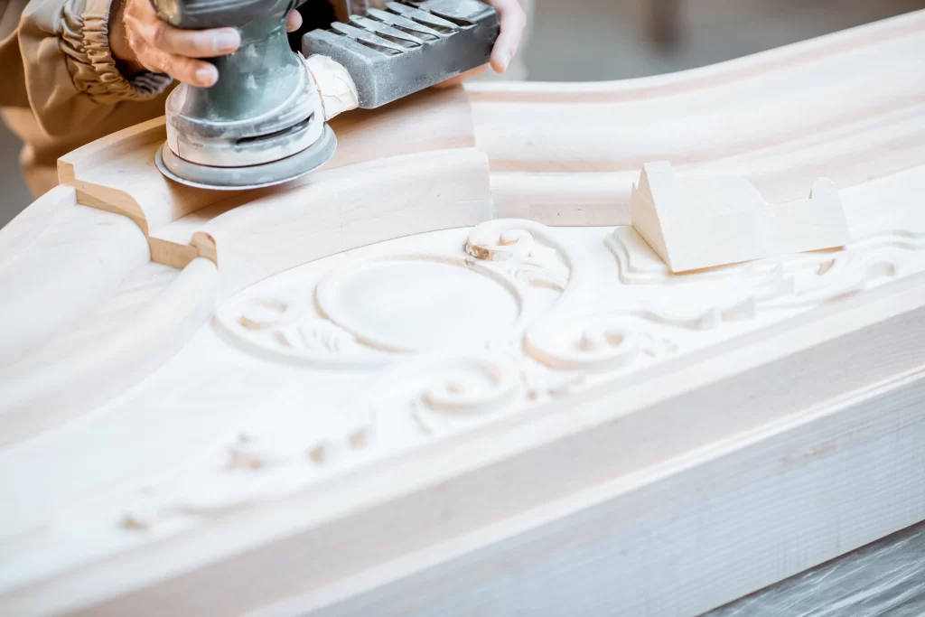 Craftsperson using orbital sander on intricately carved wooden furniture piece with decorative relief details