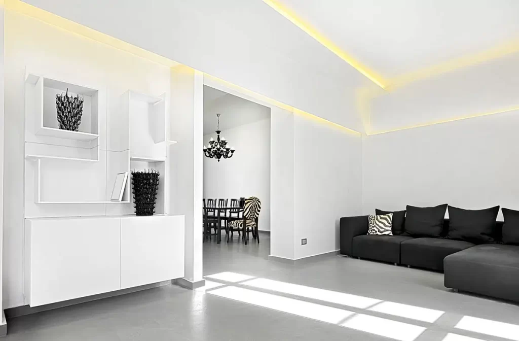 A minimalist white living room with subtle yellow cove lighting, featuring a black sofa and glimpse into a formal dining area with zebra-print chairs.
