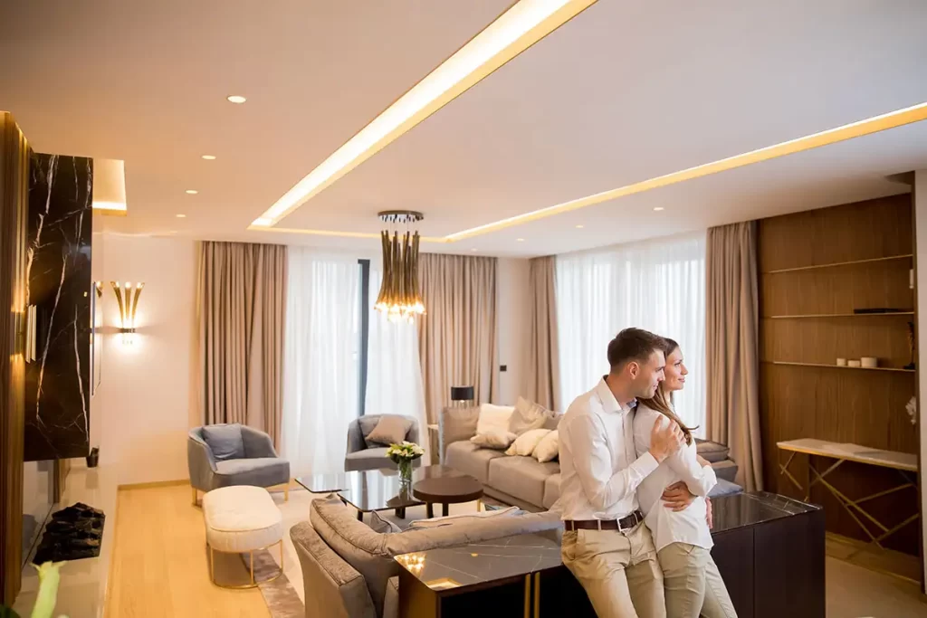 An elegant living room with warm cove lighting, light gray furniture, floor-to-ceiling curtains, and two people embracing while looking out the window.