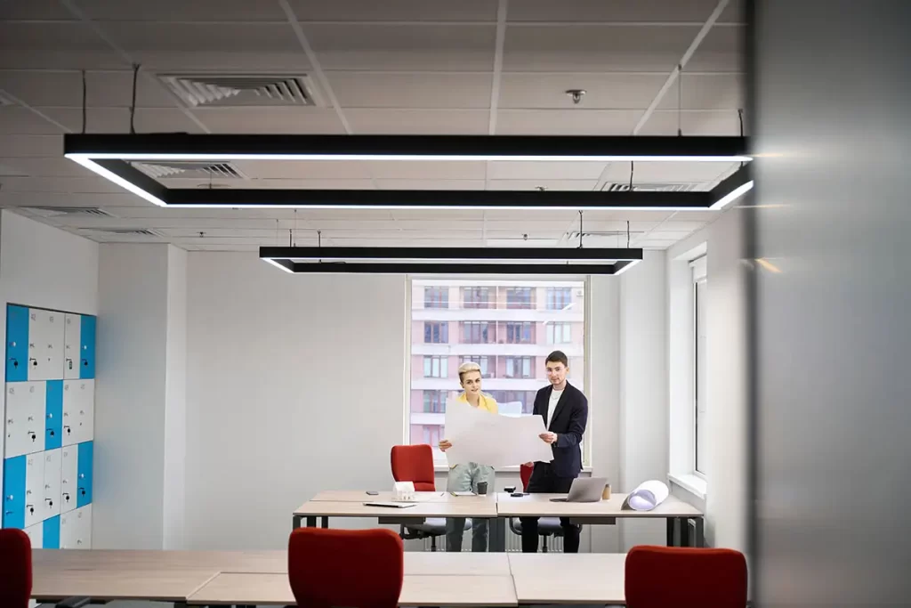  A modern office space with geometric light fixtures where two people examine documents at a desk, featuring red chairs and blue lockers.