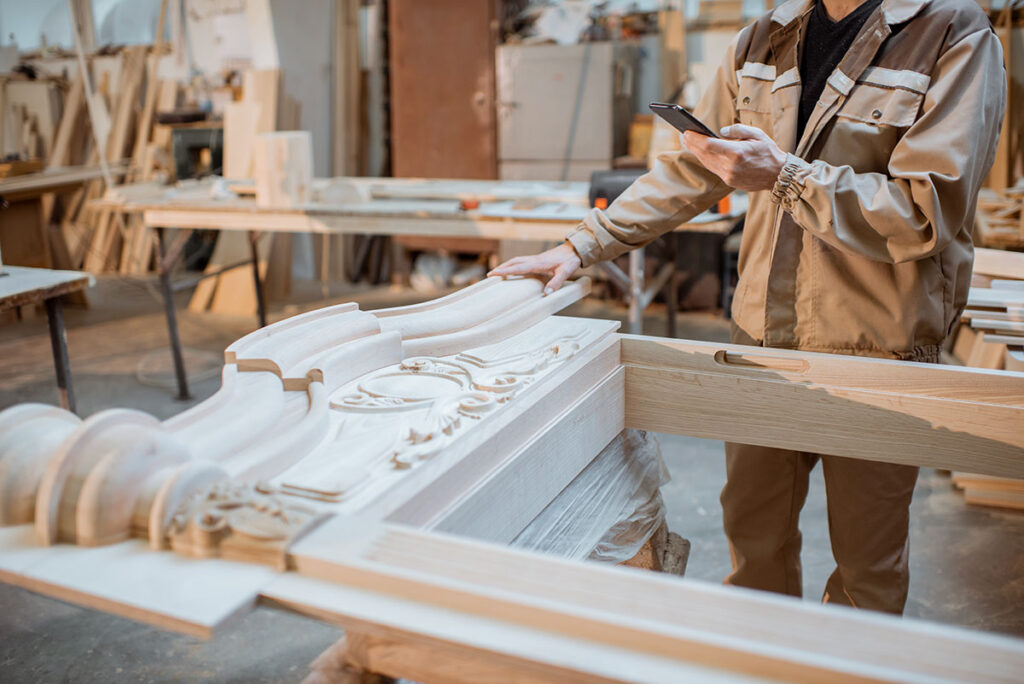 A woodworker in a beige workwear inspecting an intricately carved wooden decorative panel in a workshop while holding a smartphone, surrounded by woodworking materials and tools. 