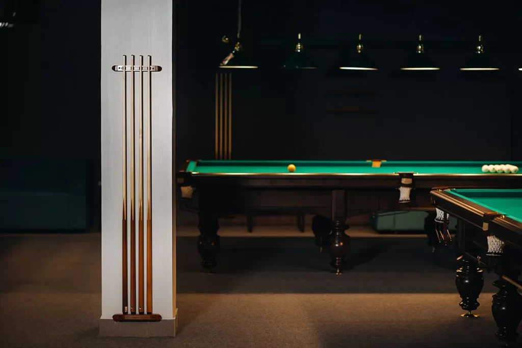 Contemporary billiards room with elegant pool tables, brass cue rack mounted on a white column, and pendant lighting