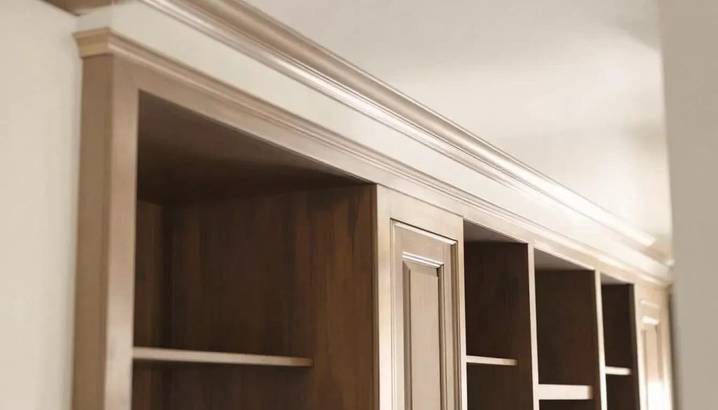 In this image, thick white crown molding elegantly frames the ceiling, enhancing the architectural detail of the room while creating a polished and sophisticated look. The clean lines of the decorative trim draw the eye upward, making the space feel taller and more cohesive, particularly around built-in bookshelves and cabinetry.